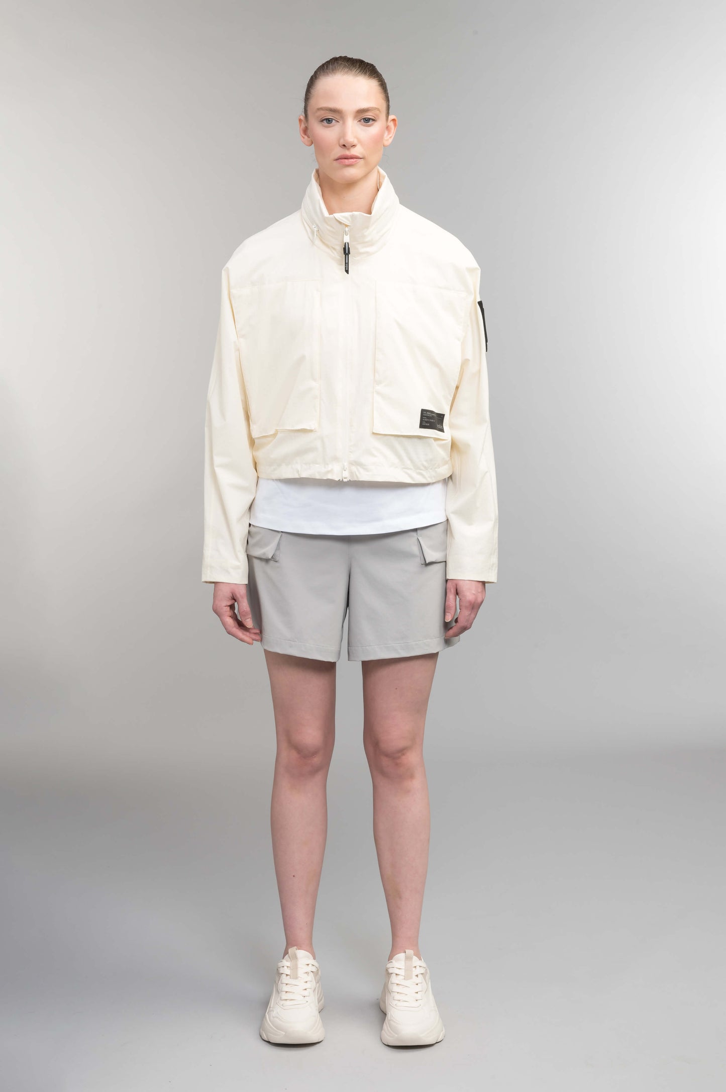 Epsie Women's Cropped Rain Jacket in Chalk crafted from premium recycled polyester ripstop with a DWR coating and taffeta nylon lining. Features a hidden packaway hood stored in the collar, two-way waterproof front zipper, seam-sealed construction, underarm zip ventilation, adjustable drawcord hem, zippered waist patch pockets, and an interior chest pocket for secure storage. Machine washable. Designed in Canada.